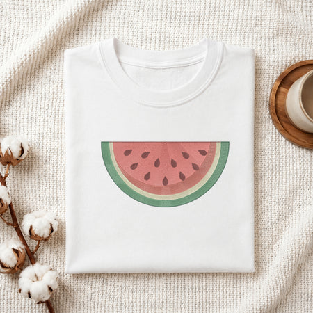 A folded white crewneck sweatshirt with a watermelon slice graphic on the front, styled on a textured cream fabric with cotton bolls and a wooden cup.