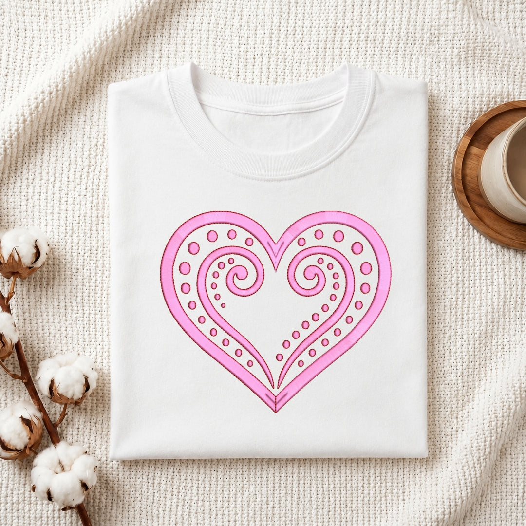 A folded white crewneck t-shirt with a pink heart graphic on the front, styled on a textured cream blanket with cotton bolls and a wooden cup in the background.