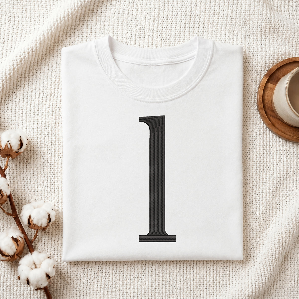 A folded white t-shirt with a large black serif number '1' printed on the front, styled in a flat lay on a textured cream fabric with cotton bolls and a wooden cup in the background.