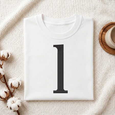A folded white t-shirt with a large black serif number '1' printed on the front, styled in a flat lay on a textured cream fabric with cotton bolls and a wooden cup in the background.