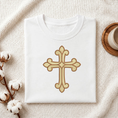 A folded white T-shirt with a large gold cross graphic on the front, styled on a textured cream fabric with cotton stems and a small cup in the background.