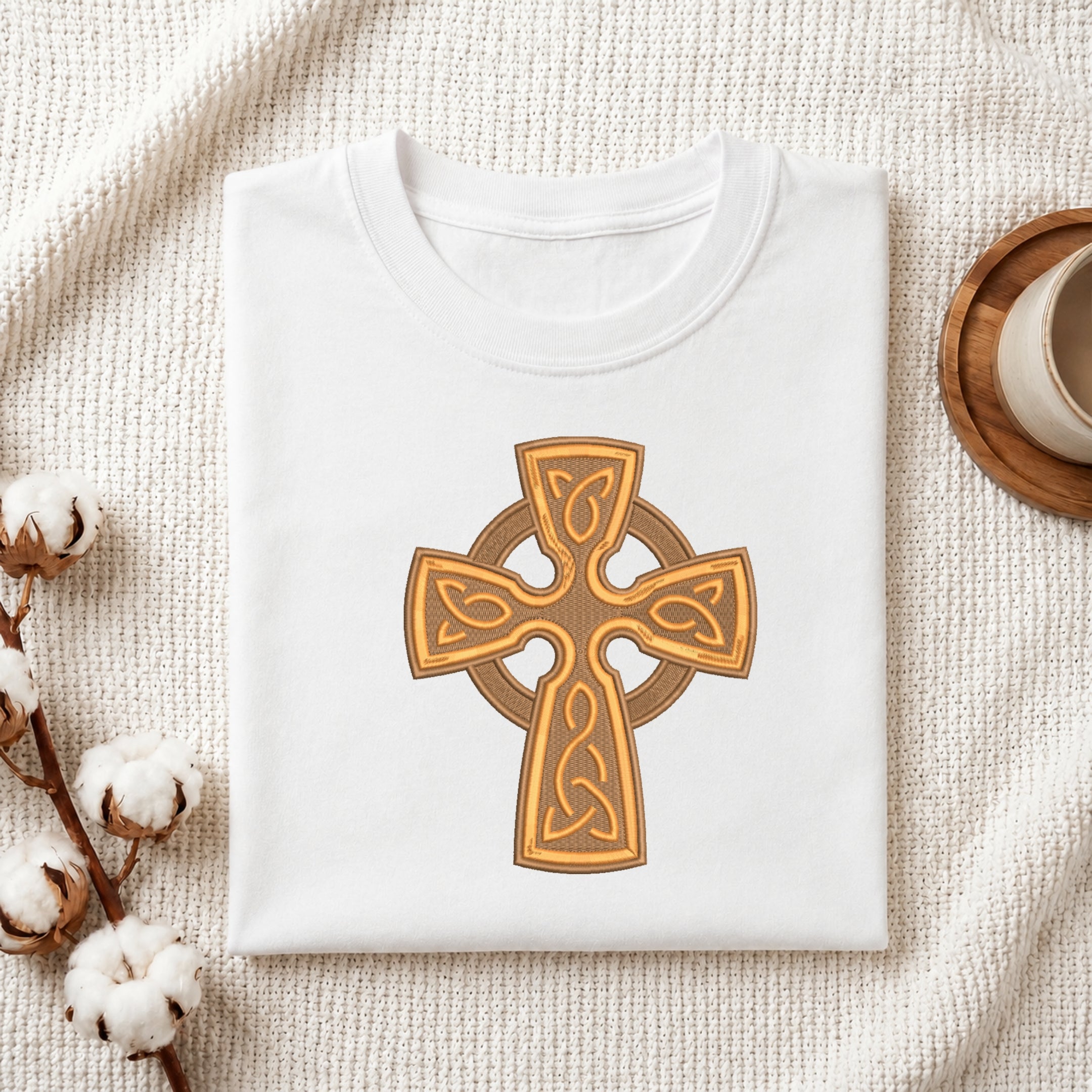 Folded white cotton t-shirt featuring an embroidered golden Celtic cross design centered on the chest.
