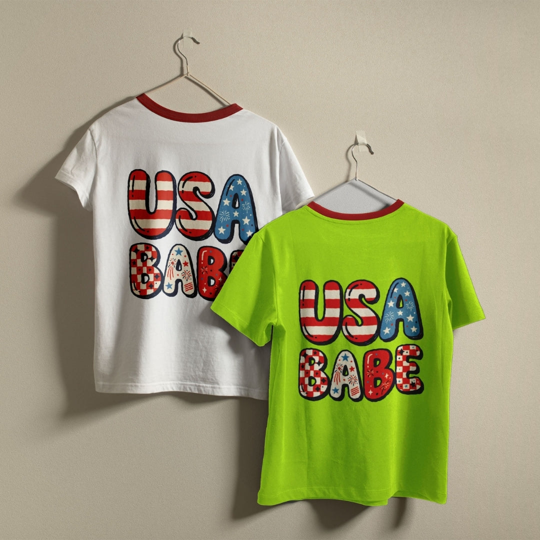 Two short-sleeve ringer T-shirts hanging on hangers against a plain beige wall. The back of a white shirt and the front of a neon green shirt both feature large 'USA BABE' text with an American flag pattern.