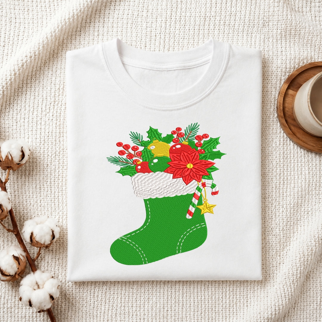 A folded white crewneck t-shirt with a Christmas stocking graphic, styled on a textured cream fabric with cotton bolls and a coffee cup.