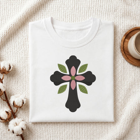 A folded white T-shirt laid flat on a textured cream fabric background, featuring a central black cross design with green leaves and a pink flower motif. Cotton bolls and a small wooden cup are placed around it as styled props.