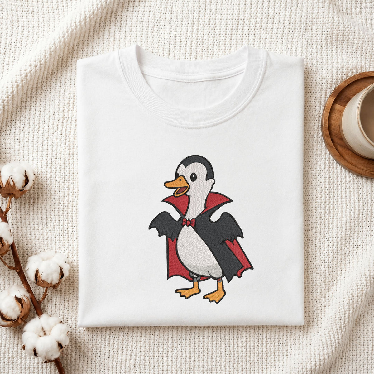 Folded white cotton t-shirt mockup featuring a cute duck in a vampire Dracula costume Halloween machine embroidery design, styled with cotton stems and a wooden cup   