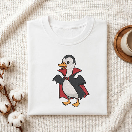 Folded white cotton t-shirt mockup featuring a cute duck in a vampire Dracula costume Halloween machine embroidery design, styled with cotton stems and a wooden cup   