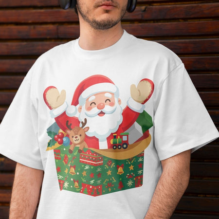A man wearing a white crewneck t-shirt with a colorful graphic of Santa Claus popping out of a Christmas gift box, set against a dark wooden background.