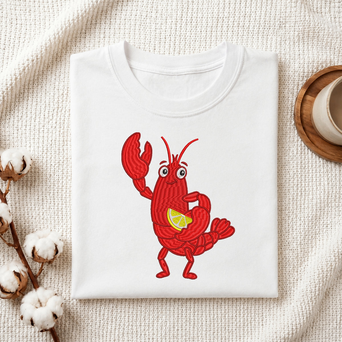 A folded white crewneck t-shirt featuring a cartoon graphic of a red lobster holding a yellow lemon slice, styled on a textured cream fabric with cotton bolls and a wooden cup in the background.