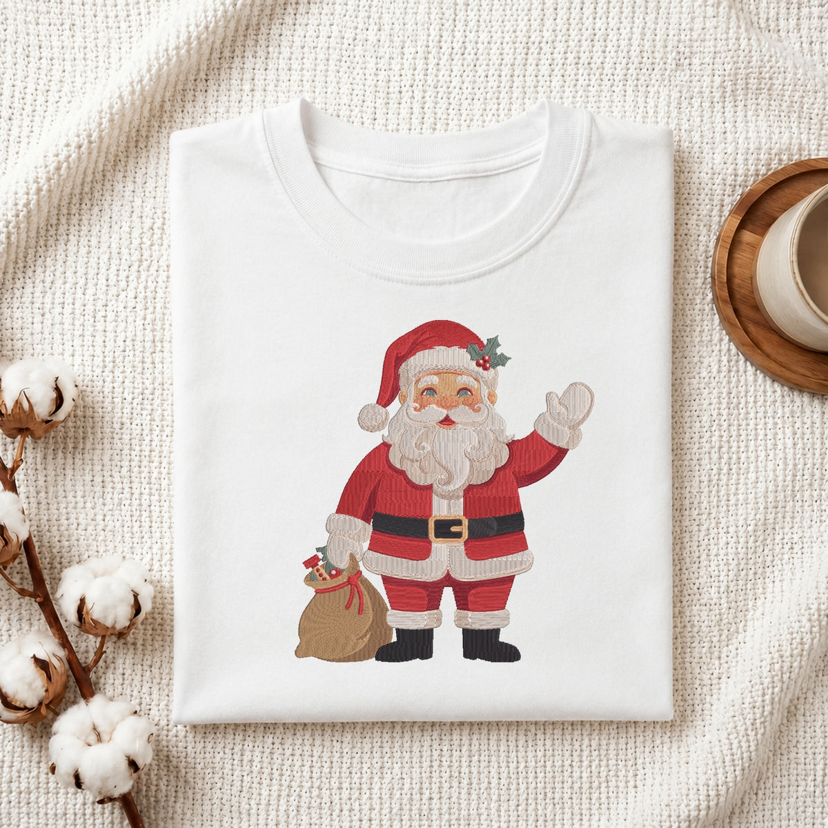 A folded white crewneck t-shirt featuring a cartoon illustration of Santa Claus waving and holding a sack, styled on a textured cream fabric with cotton bolls and a wooden cup.