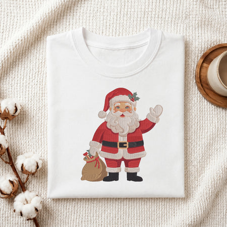 A folded white crewneck t-shirt featuring a cartoon illustration of Santa Claus waving and holding a sack, styled on a textured cream fabric with cotton bolls and a wooden cup.