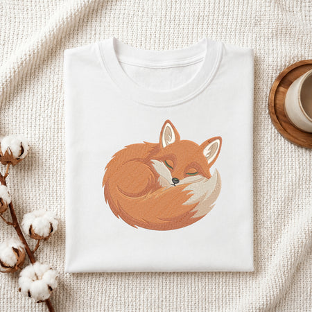 A folded white crewneck t-shirt featuring a cute cartoon illustration of a sleeping orange fox, styled on a textured cream knit blanket with cotton bolls and a wooden cup in the background.
