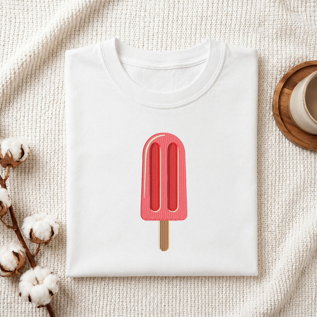 Folded white cotton t-shirt mockup featuring a pink ice cream popsicle machine embroidery design, styled with cotton stems and a wooden cup