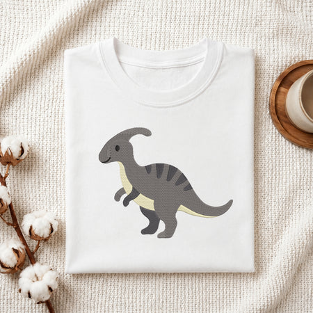 A folded white crewneck t-shirt featuring a cartoon illustration of a gray and cream Paradoxosaurus dinosaur, styled on a textured cream knit blanket with cotton bolls and a wooden cup in the background.