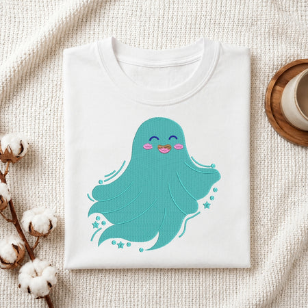 A folded white crewneck t-shirt featuring a cute embroidered-style teal octopus with pink cheeks and a small rainbow smiley, displayed on a textured cream fabric with cotton bolls and a wooden cup in the background.