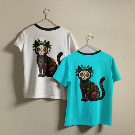 Two ringer-style t-shirts in white and bright teal hanging on a neutral wall, both displaying a detailed calavera style black cat design.
