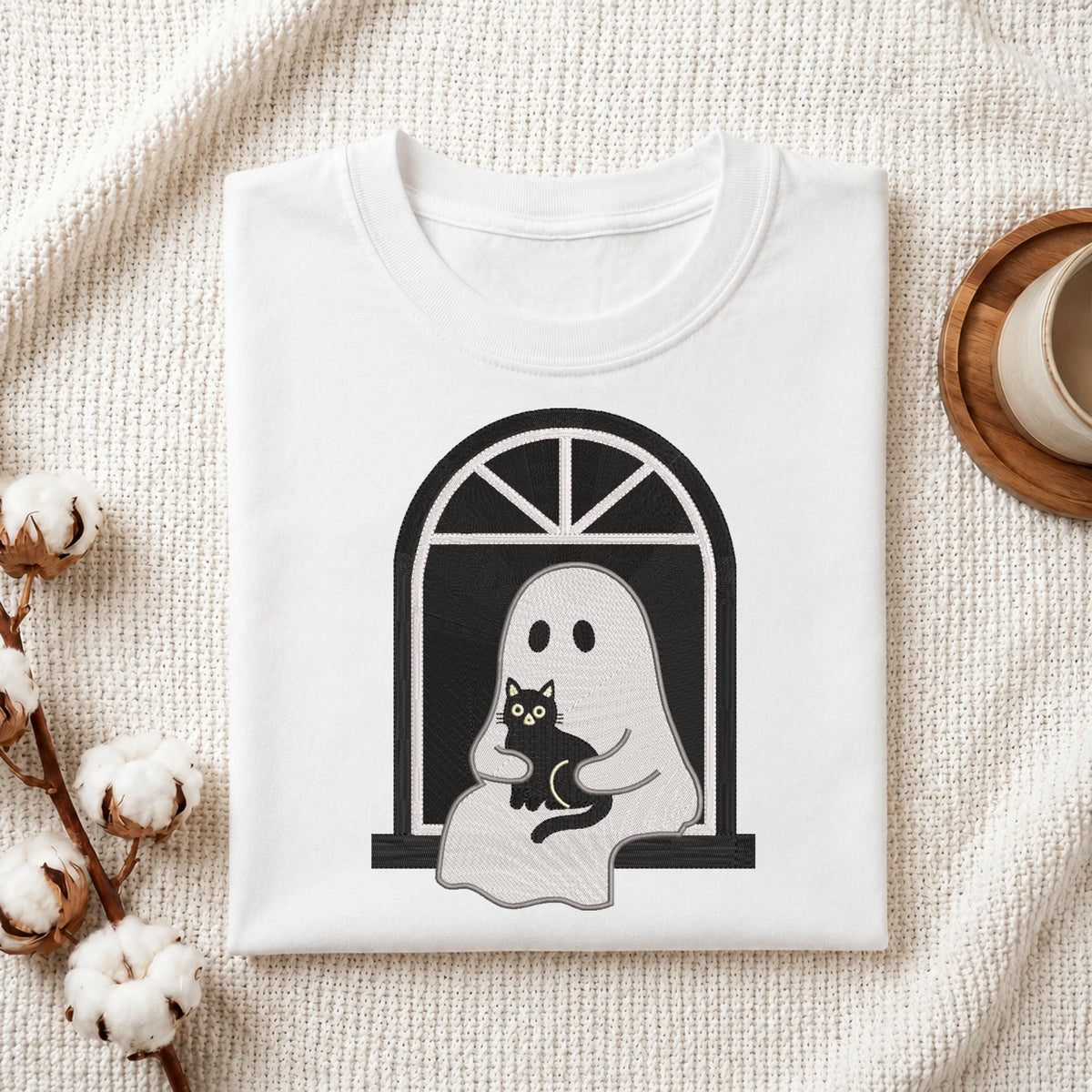 A folded white t-shirt with a black and gray graphic of a ghost holding a black cat, styled on a textured cream fabric with cotton bolls and a wooden cup.
