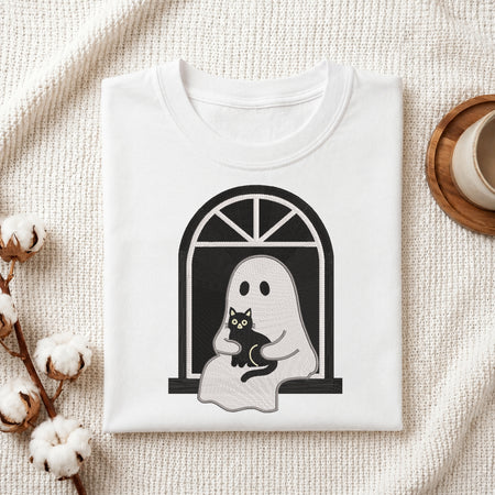 A folded white t-shirt with a black and gray graphic of a ghost holding a black cat, styled on a textured cream fabric with cotton bolls and a wooden cup.