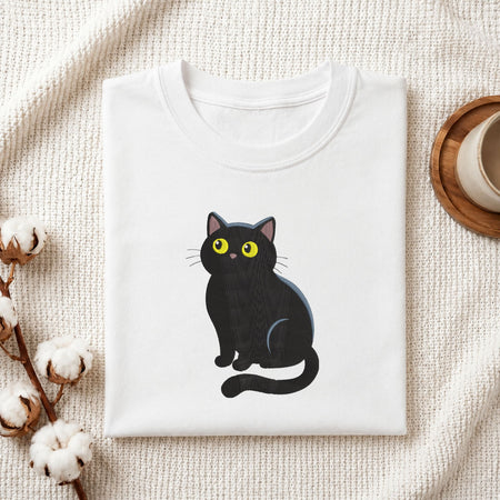 Folded white cotton t-shirt mockup featuring a cute black cat with yellow eyes machine embroidery design, styled with cotton stems and a wooden cup.