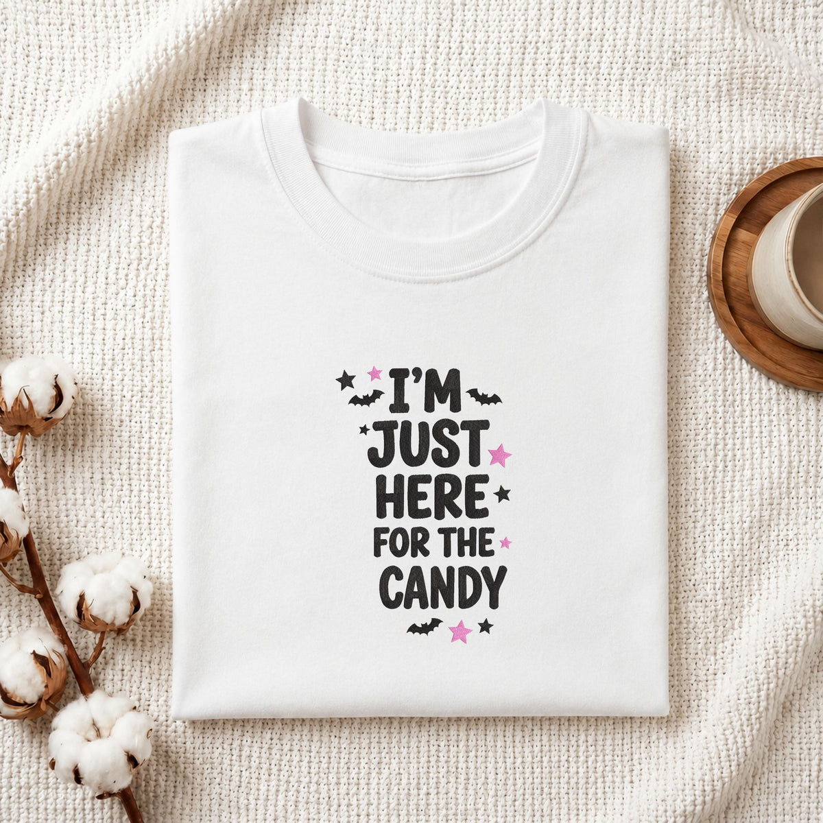 Folded white cotton t-shirt mockup featuring a "I'm Just Here for the Candy" Halloween machine embroidery design, styled with cotton stems and a wooden cup