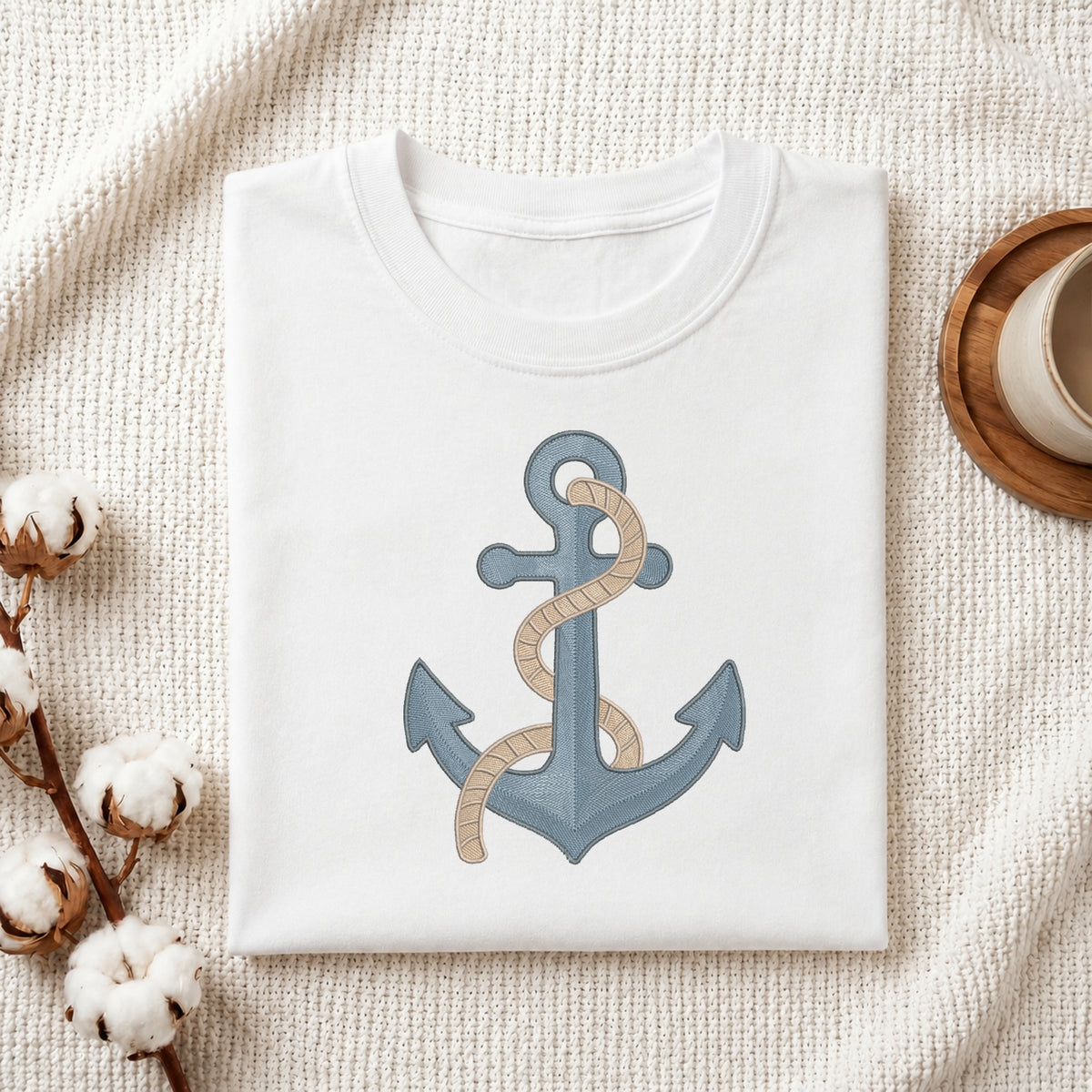 A folded white crewneck t-shirt featuring a graphic of a blue anchor wrapped with beige rope, styled on a textured cream fabric with cotton bolls and a wooden cup.