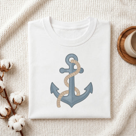 A folded white crewneck t-shirt featuring a graphic of a blue anchor wrapped with beige rope, styled on a textured cream fabric with cotton bolls and a wooden cup.