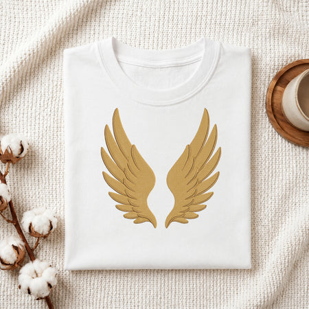 A folded white crewneck t-shirt with a gold embroidered angel wings design on the front, styled on a textured cream fabric with cotton bolls and a wooden cup.