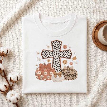 A folded white t-shirt with a fall-themed graphic print featuring a cross and pumpkins, styled on a textured cream blanket with cotton stems and a mug.