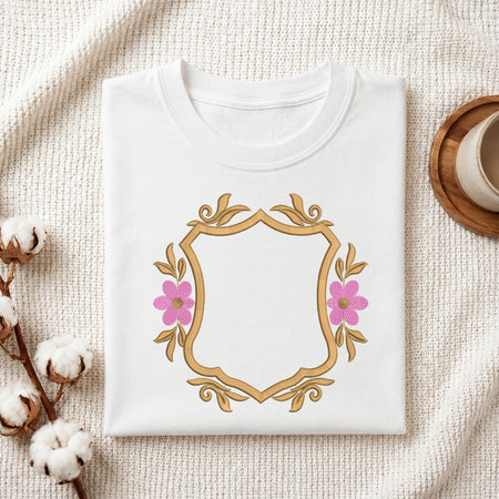 A folded white t-shirt with a gold ornate frame design and pink flowers on the front, displayed on a textured cream fabric alongside cotton bolls and a wooden cup.