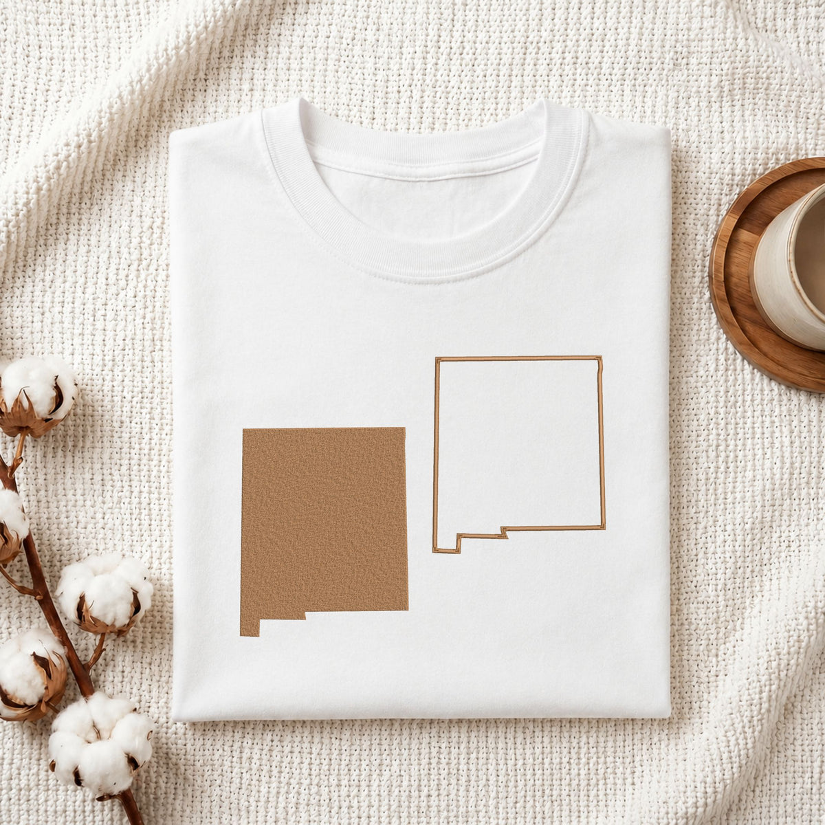 A folded white crewneck t-shirt with a graphic of the state of New Mexico in two designs (solid brown and outlined) on the front, styled on a textured cream blanket with cotton bolls and a coffee cup.