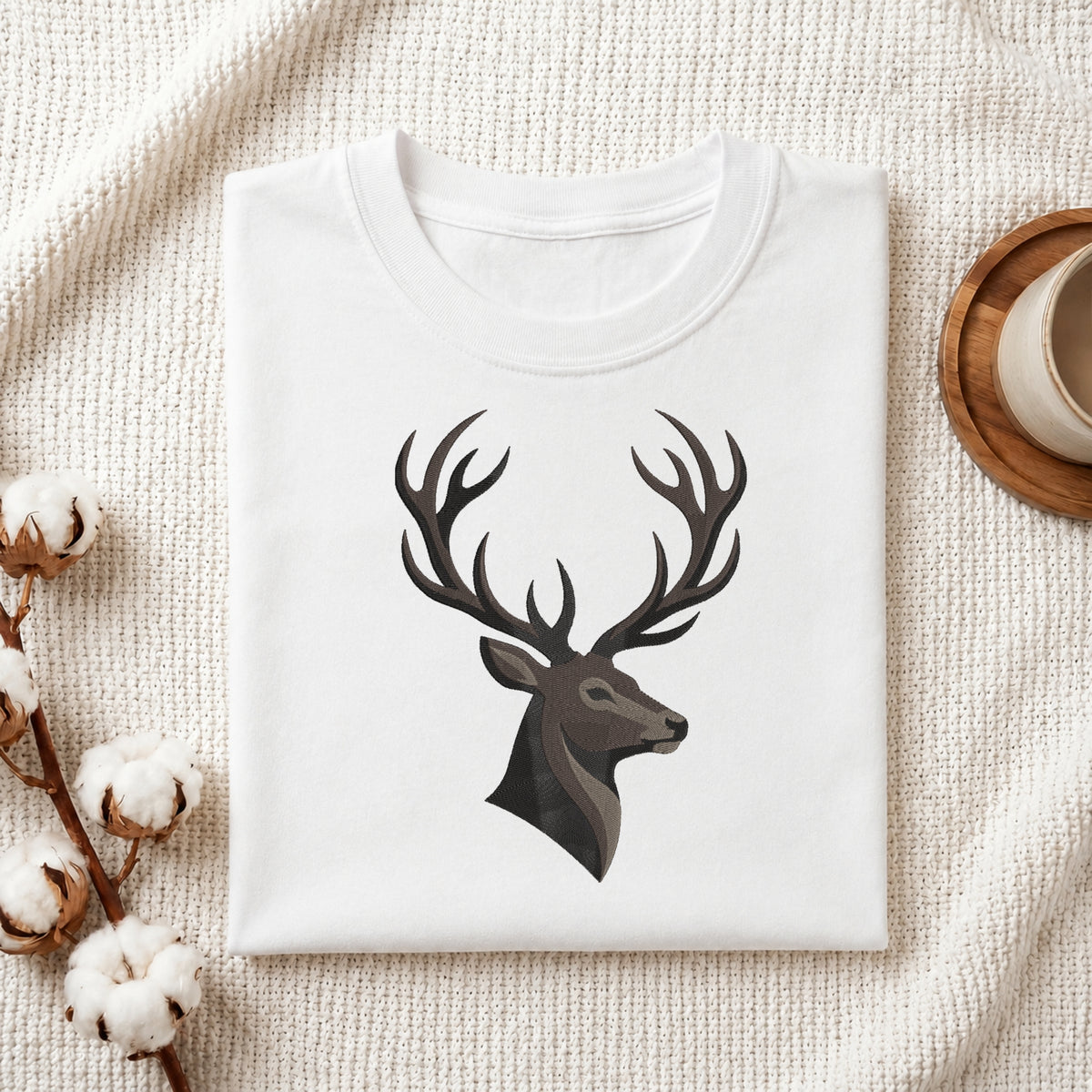 A folded white crewneck t-shirt with a dark gray stag head graphic on the chest, styled on a textured cream knit blanket with cotton bolls and a wooden cup in the background.