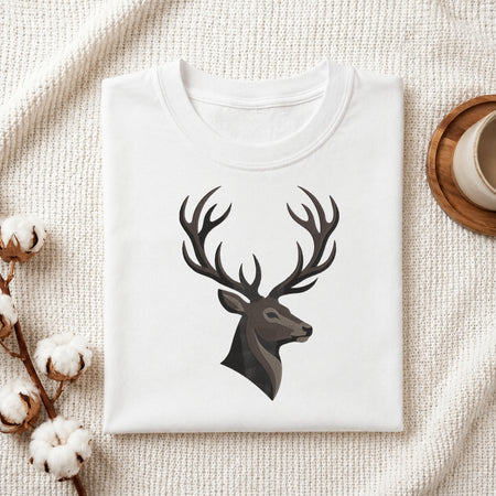 A folded white crewneck t-shirt with a dark gray stag head graphic on the chest, styled on a textured cream knit blanket with cotton bolls and a wooden cup in the background.