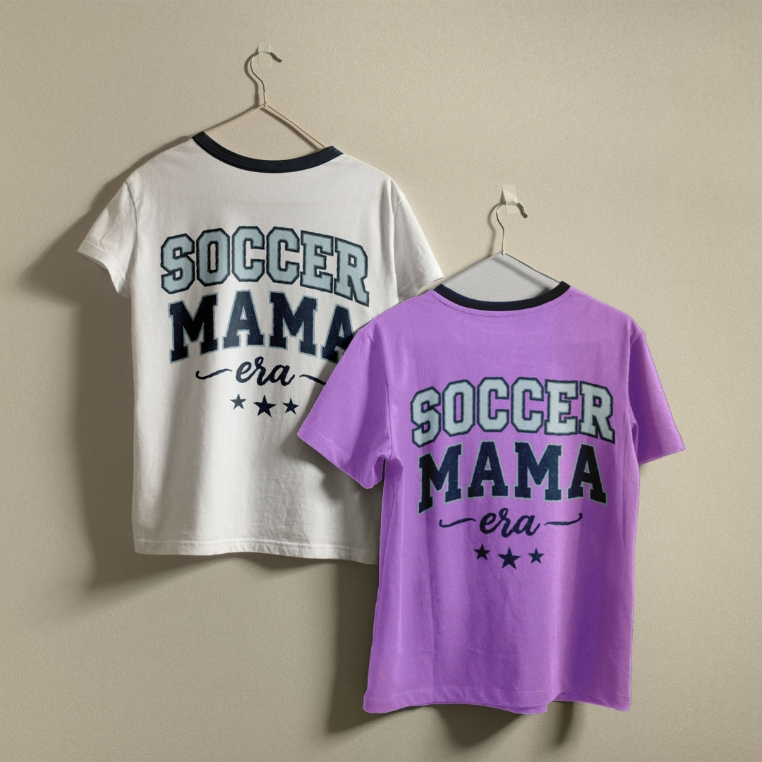 Two t-shirts hanging on a wall, one white and one purple, both featuring the retro "Soccer Mama Era" sports graphic design.