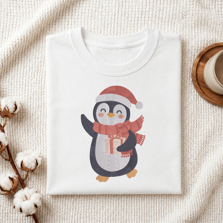 A folded white crewneck t-shirt featuring a cute cartoon penguin wearing a red Santa hat and matching red scarf, holding a small gift box. The t-shirt is styled in a flat-lay arrangement on a textured cream knit blanket, with cotton bolls on the left and a wooden cup on the right.