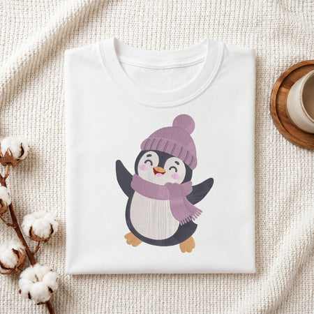 A folded white crewneck t-shirt featuring a cute cartoon penguin wearing a purple knit hat and matching scarf, styled on a textured cream knit blanket with cotton bolls and a wooden cup in the background.