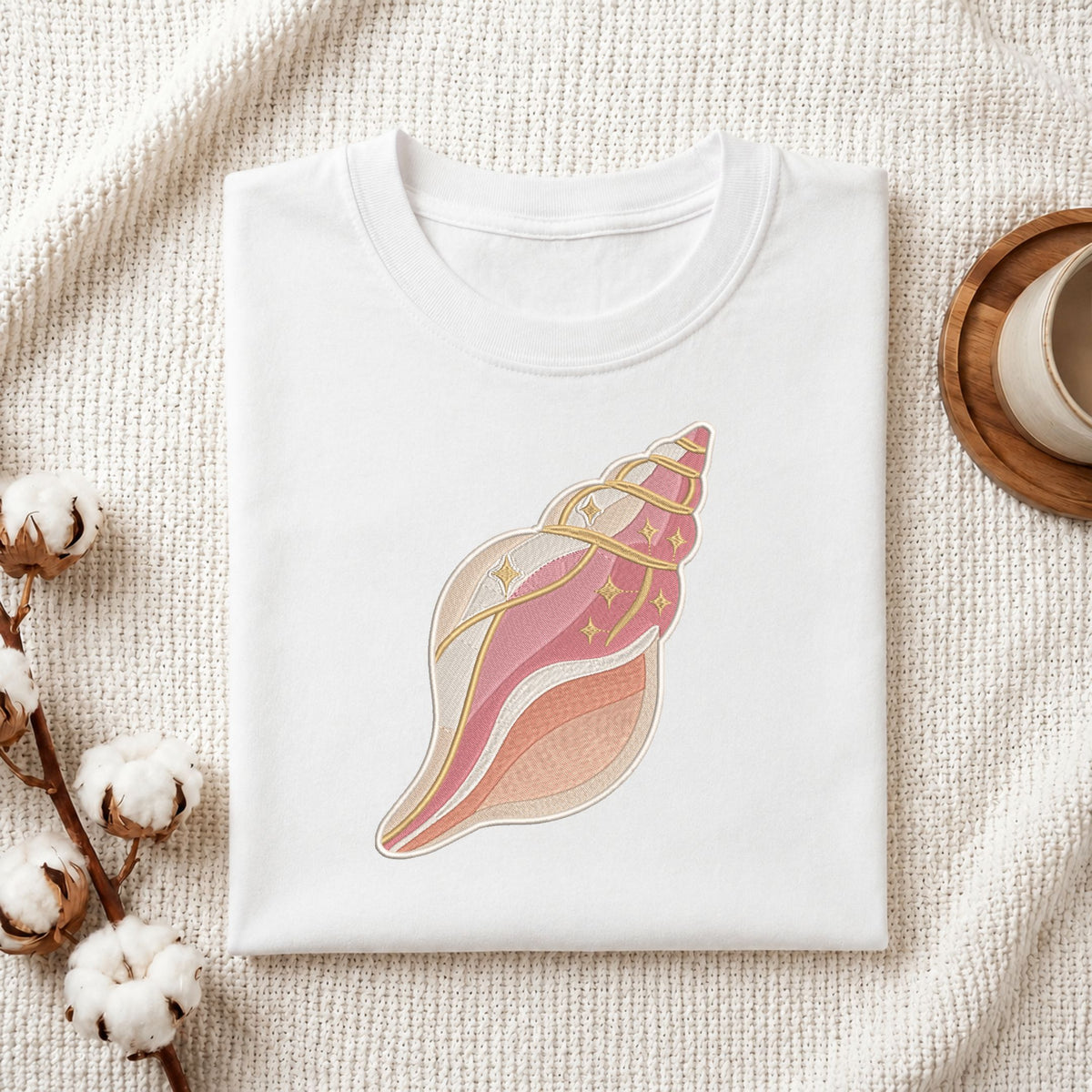 Folded white cotton t-shirt mockup featuring a pink and beige conch seashell machine embroidery design, styled with cotton stems and a wooden cup.