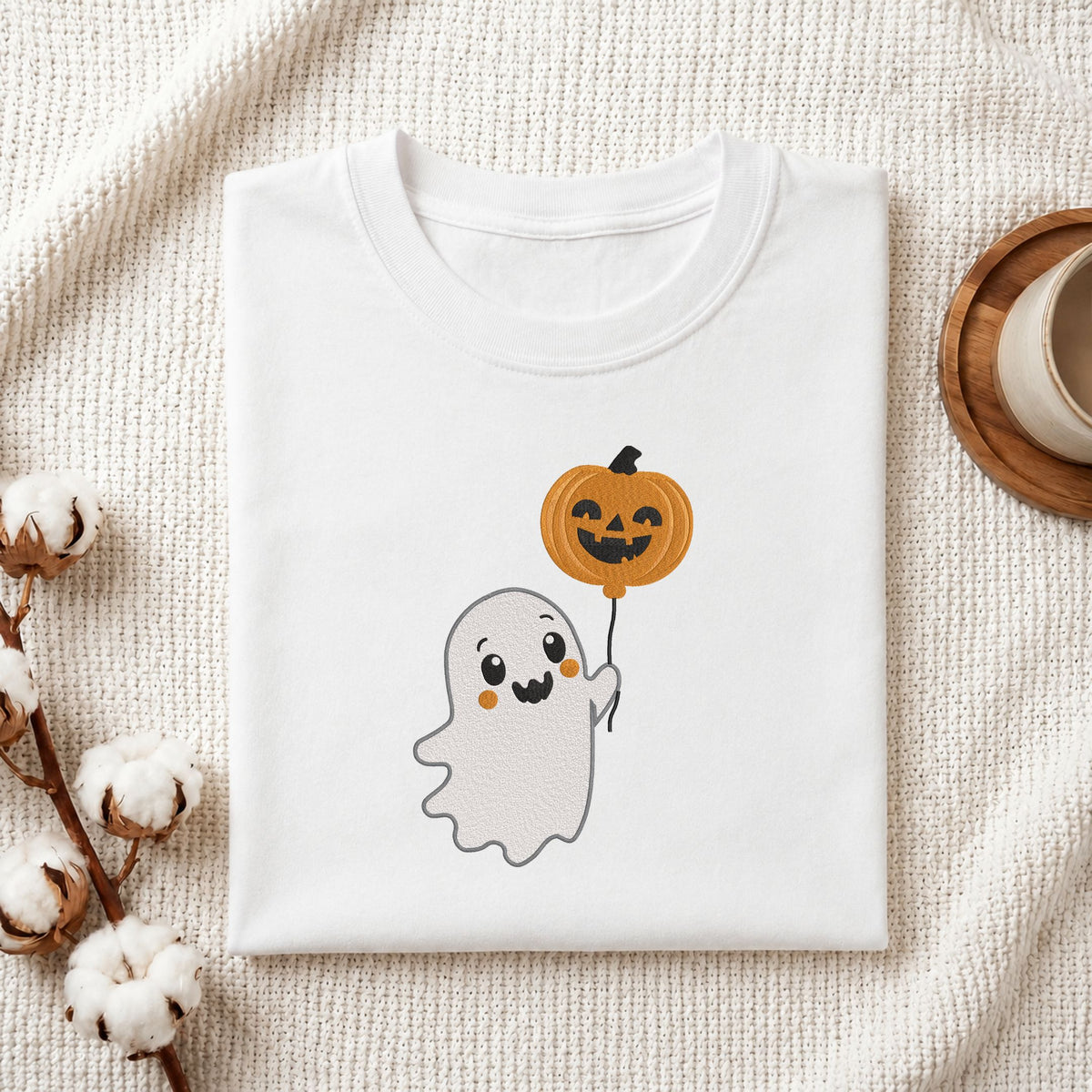Folded white cotton t-shirt mockup featuring a cute ghost machine embroidery design, styled with cotton stems and a wooden cup