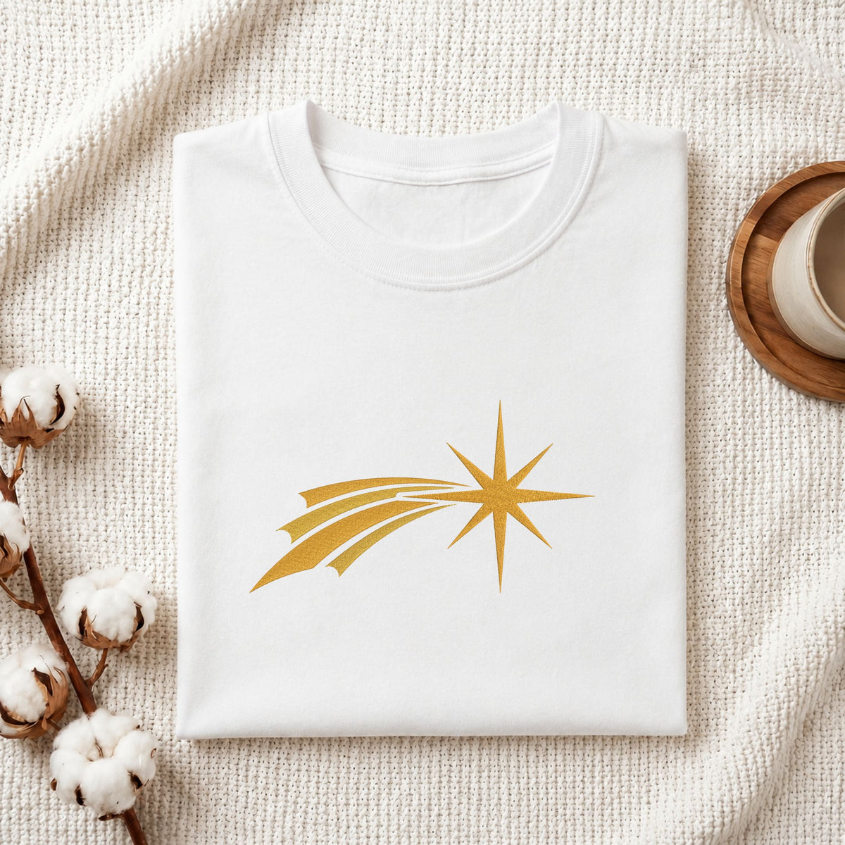 Folded white cotton t-shirt mockup featuring a golden comet star machine embroidery design, styled with cotton stems and a wooden cup
