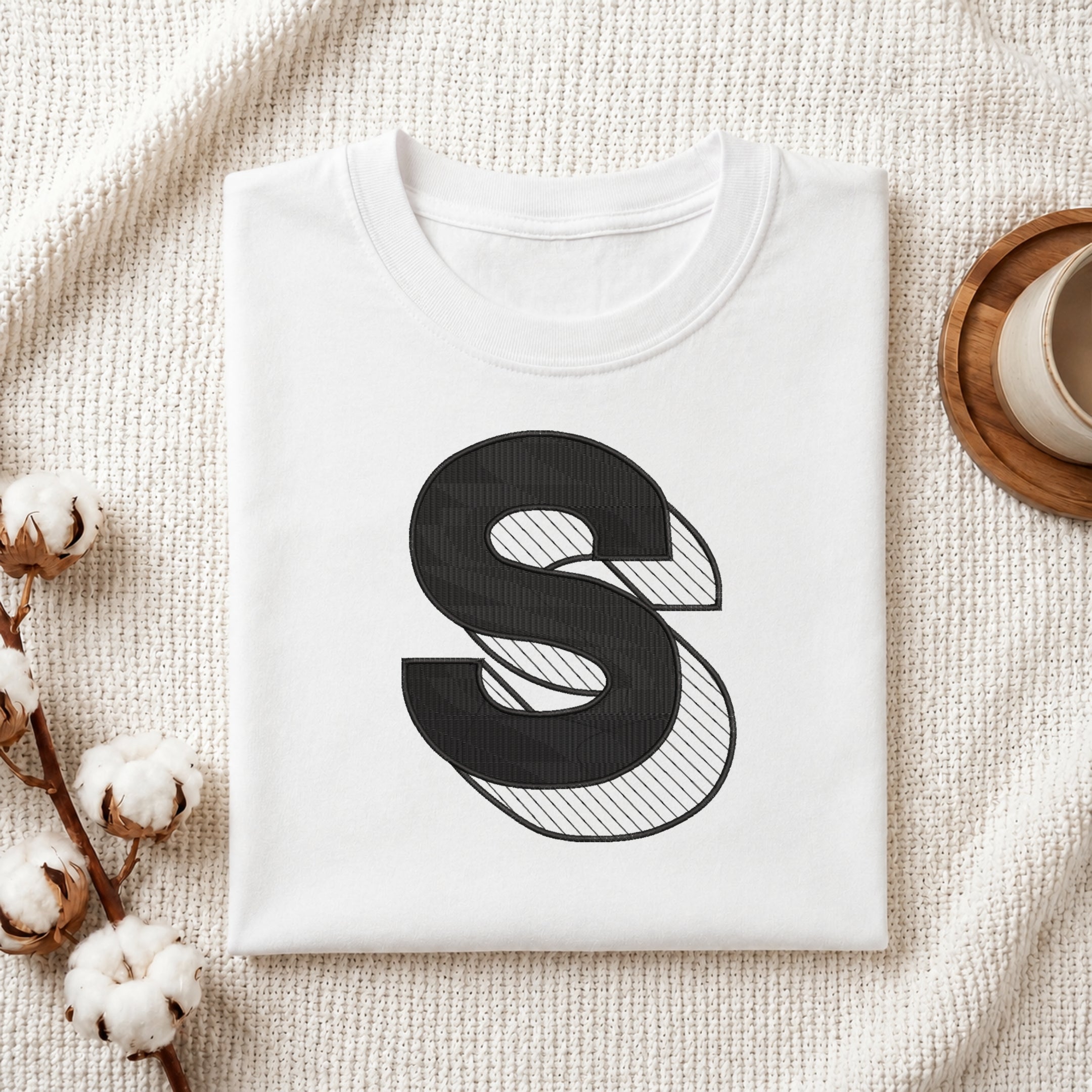 A folded white crewneck t-shirt with a large black stylized letter 'S' graphic on the front, styled on a textured cream knit blanket with cotton stems and a wooden cup in the background.