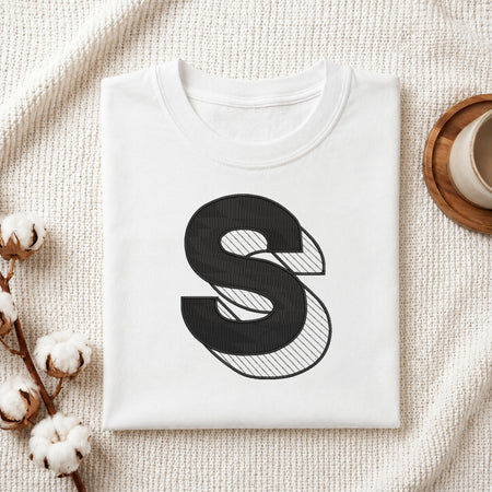 A folded white crewneck t-shirt with a large black stylized letter 'S' graphic on the front, styled on a textured cream knit blanket with cotton stems and a wooden cup in the background.
