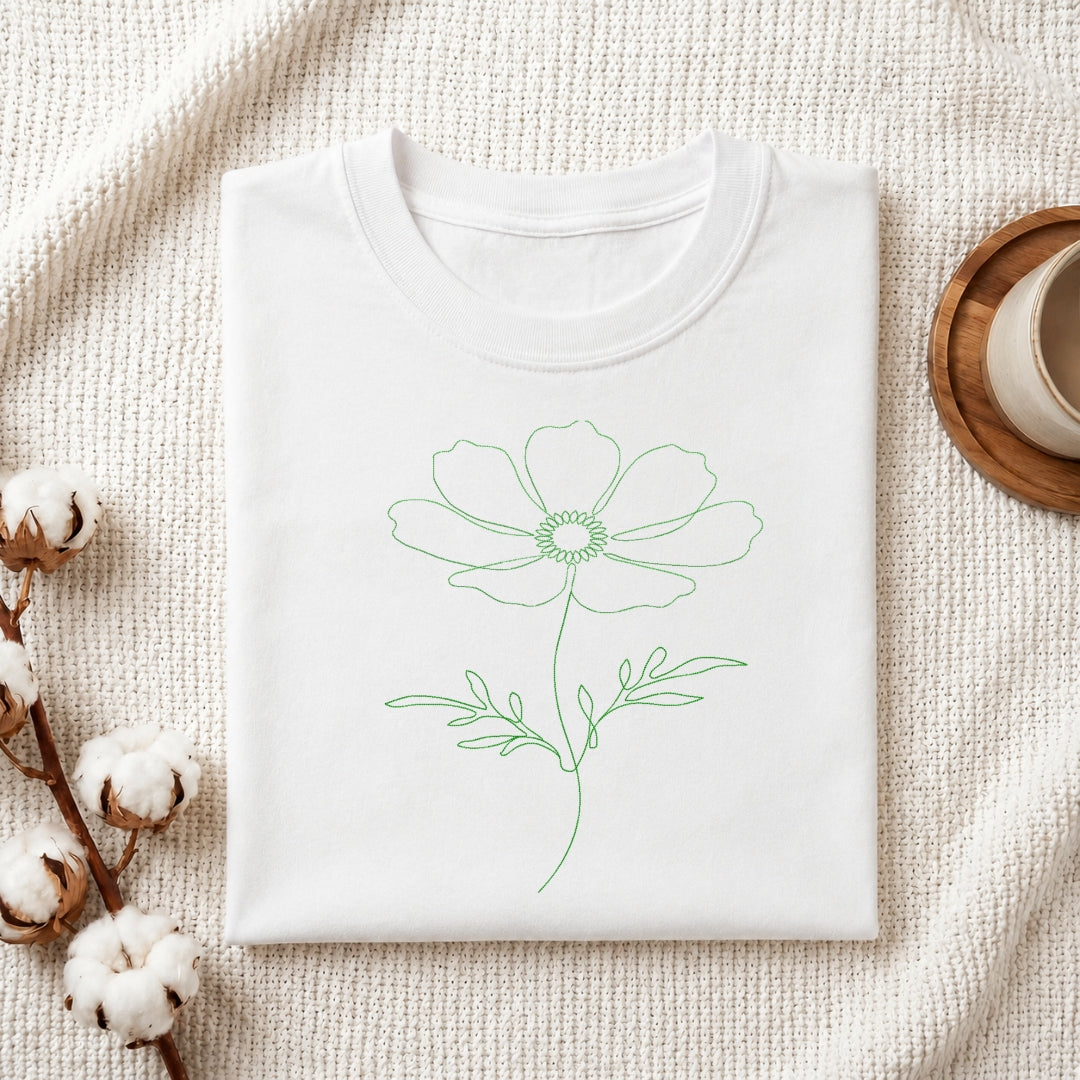 A folded white crewneck t-shirt with a simple green line art flower design on the front, styled on a textured cream fabric with cotton bolls and a wooden cup in the background.