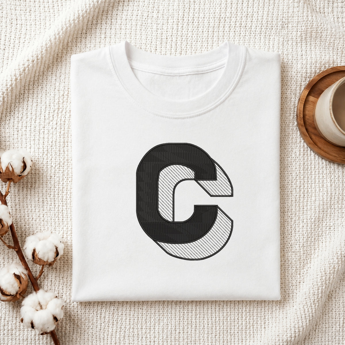 A folded white crewneck t-shirt with a large black graphic letter 'C' on the front, styled on a textured cream knit blanket with cotton bolls and a wooden cup in the background.