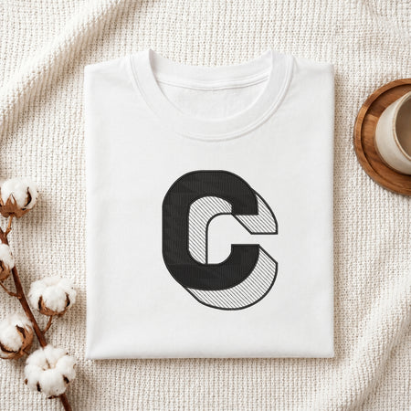 A folded white crewneck t-shirt with a large black graphic letter 'C' on the front, styled on a textured cream knit blanket with cotton bolls and a wooden cup in the background.
