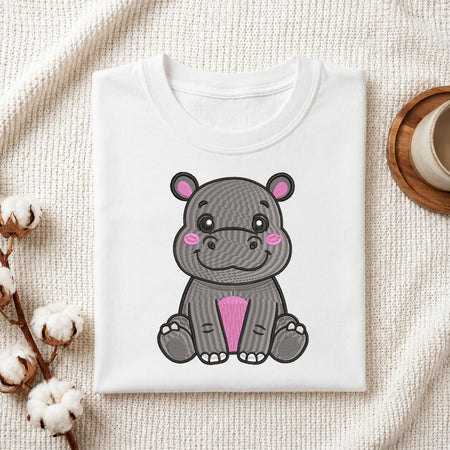 A folded white T-shirt with a cute cartoon hippo graphic on the front, styled on a textured cream fabric with cotton stems and a small wooden cup in the background.