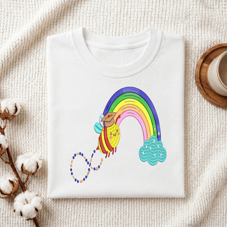 A folded white crewneck t-shirt with a colorful cartoon bee and rainbow graphic, styled on a textured cream fabric with cotton bolls and a wooden cup in the background.
