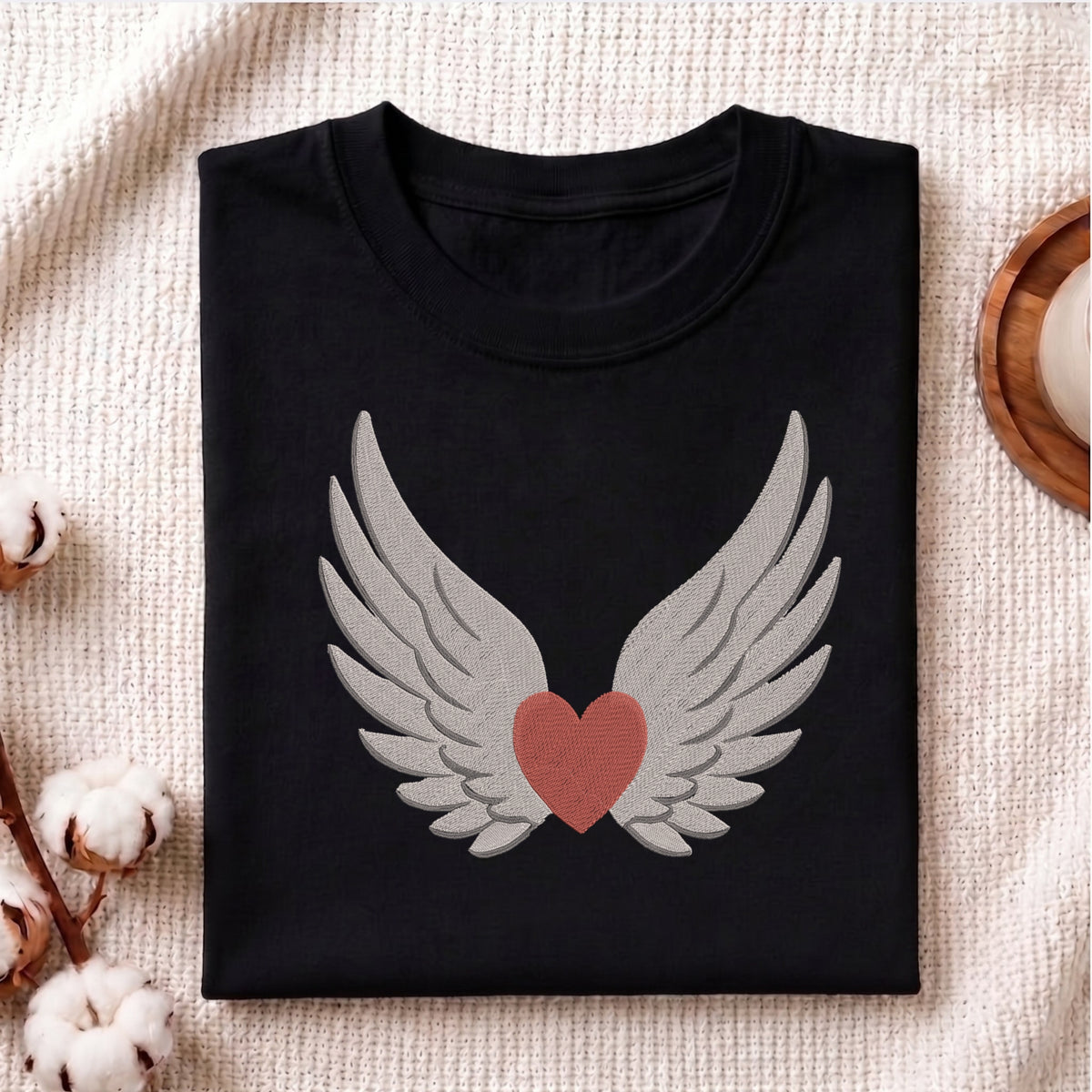 A folded black crewneck t-shirt featuring a graphic of white angel wings enclosing a dusty rose red heart, displayed on a textured beige fabric background with decorative cotton bolls.
