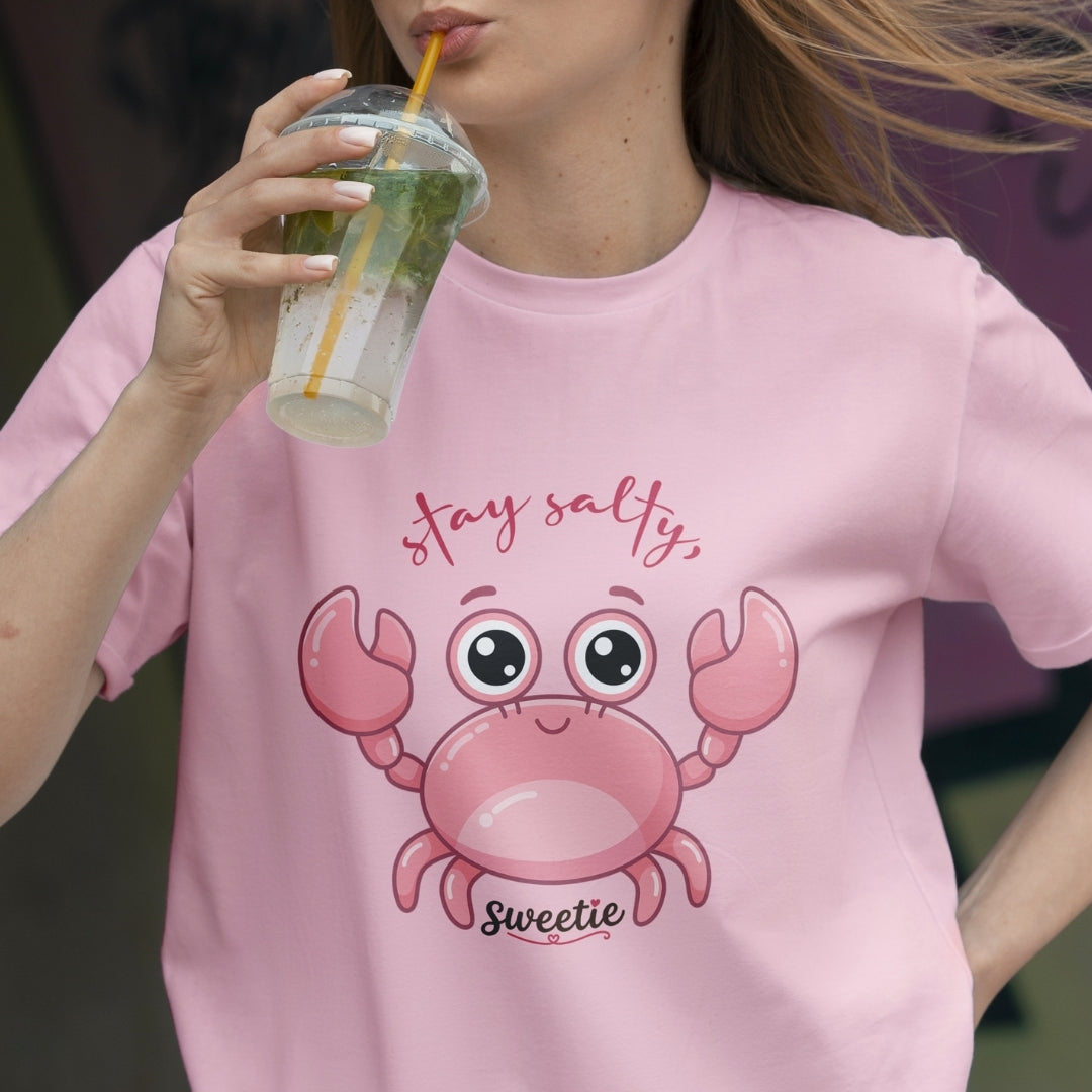 Person wearing a pink t-shirt with a cartoon crab design and text, holding a drink.