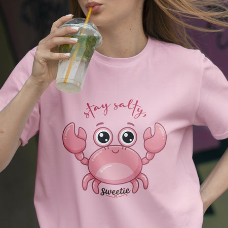 Person wearing a pink t-shirt with a cartoon crab design and text, holding a drink.