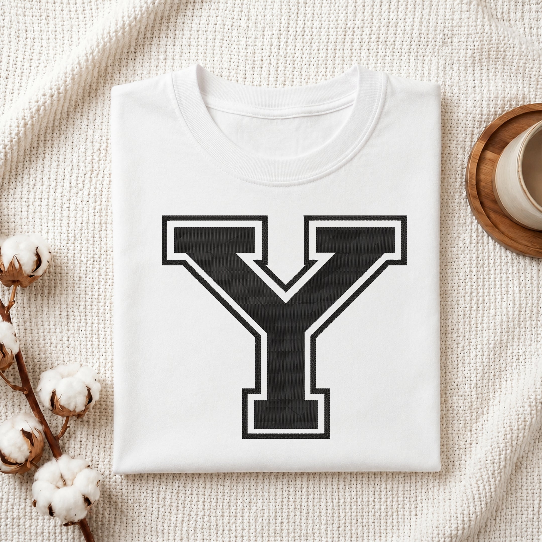 A folded white T-shirt with a large black block letter Y outlined in white on the front, styled on a textured cream fabric with cotton stems and a small cup at the side.