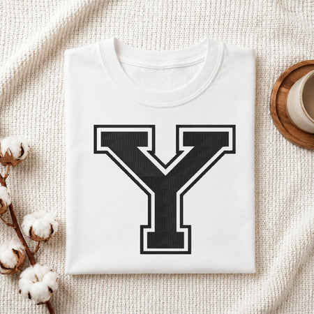 A folded white T-shirt with a large black block letter Y outlined in white on the front, styled on a textured cream fabric with cotton stems and a small cup at the side.